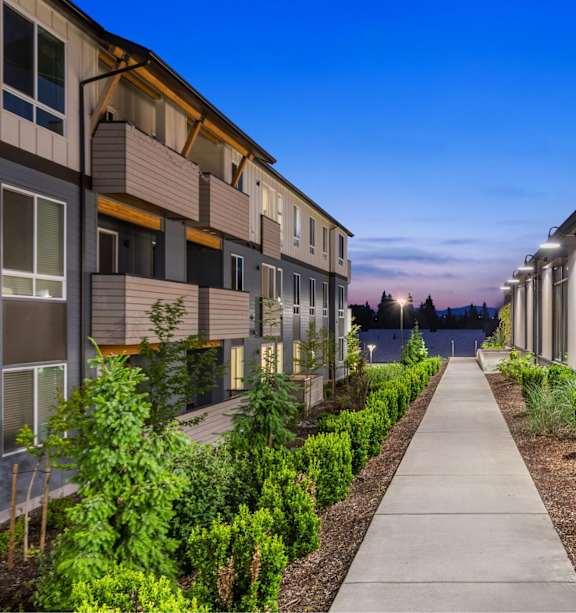 A long walkway separates apartment buildings with green shrubbery on either side.