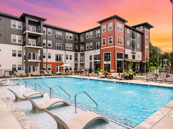 A large swimming pool is surrounded by lounge chairs in front of apartment buildings.