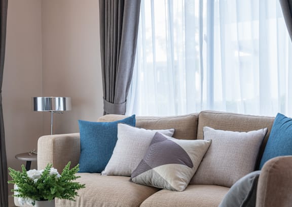 A beige couch with blue and white pillows sits in front of a window with sheer curtains.