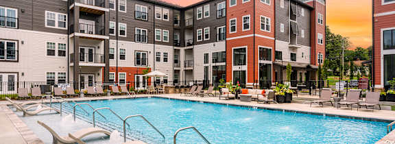 A large swimming pool is surrounded by lounge chairs in front of apartment buildings.