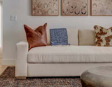 A white couch with a brown pillow and a blue blanket is in the foreground of a room with a patterned rug and a round coffee table.