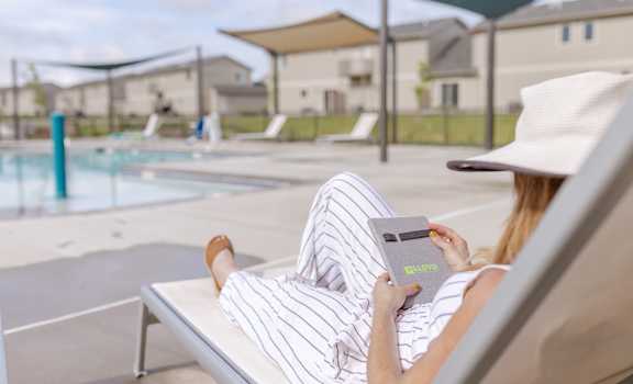 A woman in a striped dress and hat is reading a book by the pool.