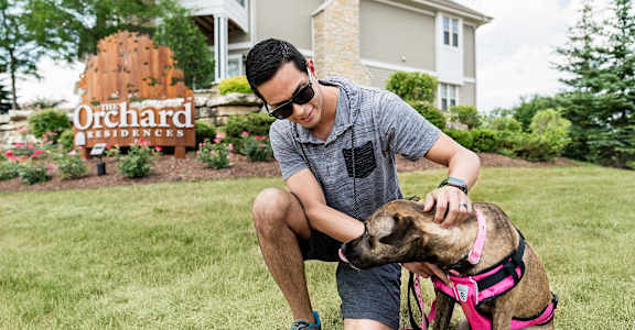 A man is petting a dog in front of a sign that says "Orchard Residences.".