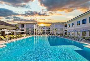 A large swimming pool surrounded by lounge chairs and buildings in the background.