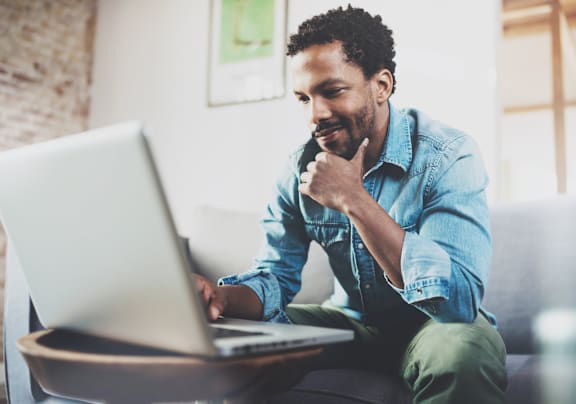 A man in a denim shirt is sitting on a couch using a laptop. at Vintage Edge Nashville Apartments, Tennessee, 37211