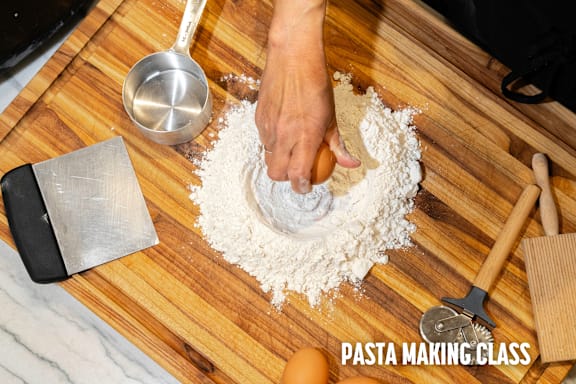 A person is kneading dough on a wooden surface with a spoon and a spatula nearby.