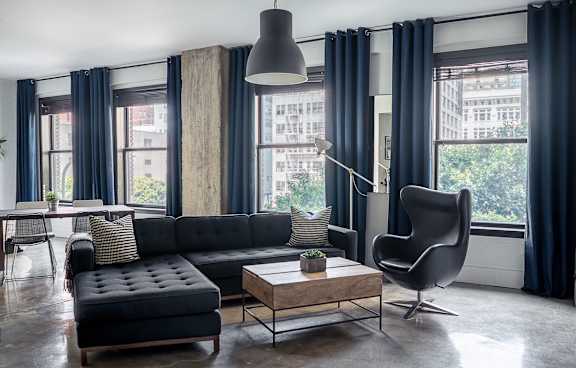 A black couch with a wooden coffee table in front of a window with a chair and a floor lamp.