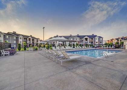 A large outdoor swimming pool surrounded by lounge chairs and buildings in the background.