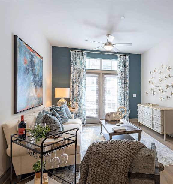A living room with a sofa, a coffee table, a painting, a ceiling fan, and a bedroom in the background.