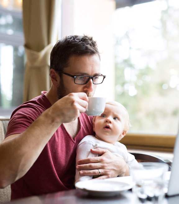 A man holding a baby while drinking from a cup in front of a laptop.at Mainline North Apartments - Santa Clara, CA, Santa Clara California