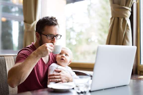 A man holding a baby while drinking from a cup in front of a laptop.