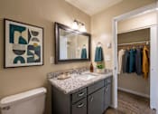 Thumbnail 5 of 8 - A bathroom with a white toilet, a sink with a marble countertop, a mirror above the sink, a wooden floor, and a painting on the wall.