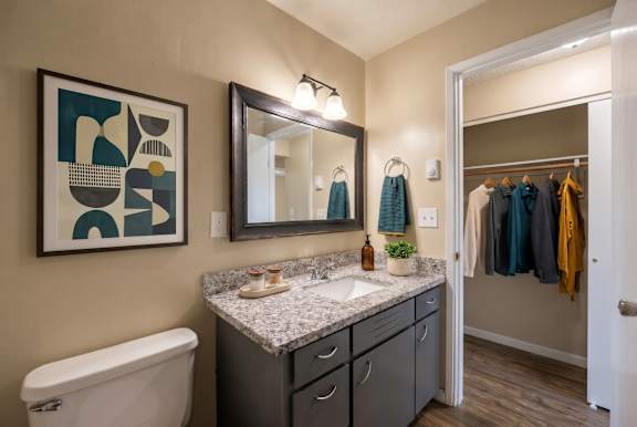 A bathroom with a white toilet, a sink with a marble countertop, a mirror above the sink, a wooden floor, and a painting on the wall.