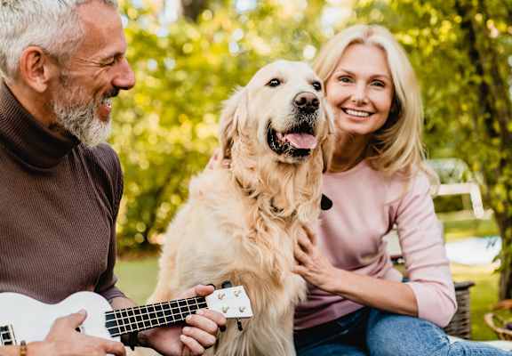 A man and woman are sitting in a park with a dog, and the man is playing a guitar.