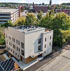 A modern building with a flat roof is surrounded by trees and other buildings.