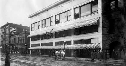 A black and white photo of a street with a horse and carriage in front of a building.