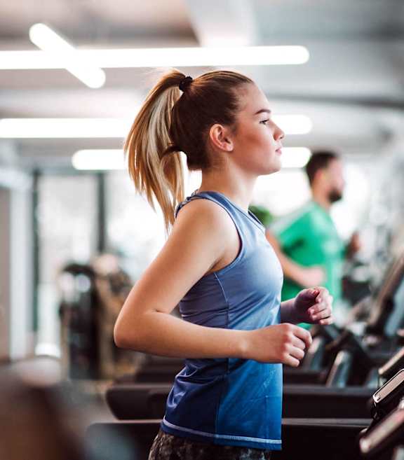 A woman in a blue top and grey shorts is running on a treadmill in a gym.