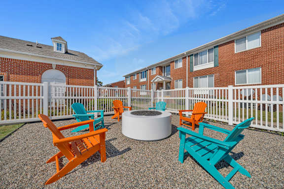 A white fence surrounds a fire pit with orange and blue chairs in front of a brick building.