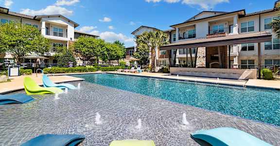 A pool with blue and green loungers in front of apartment buildings.