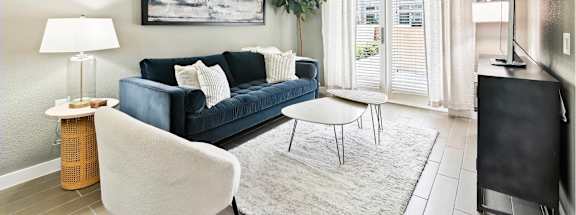 A living room with a blue couch, a white chair, a small table, a ceiling fan, and a painting on the wall.