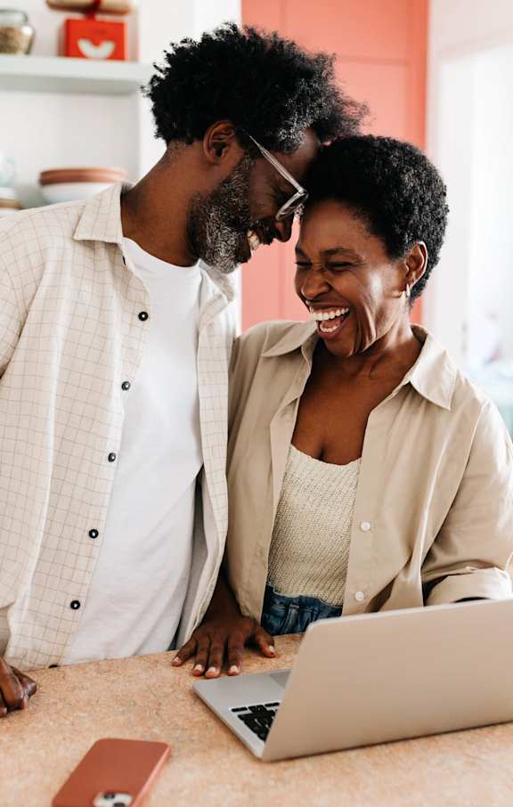 A man and woman are standing close to each other in a kitchen, looking at a laptop screen.