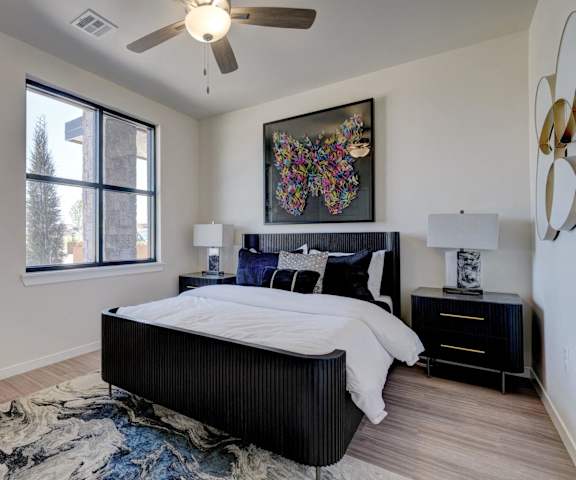 A spacious bedroom with a queen-size bed with a bedspread, a dresser, wall art, lamps, decorations, overhead lighting with a ceiling fan, large window, and nightstands with lamps at the Encore Apartments. 