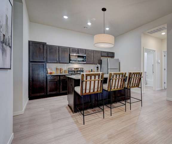 A large, modern kitchen with stainless steel appliances, bar counter with barstools, kitchen utensils, marble countertops with lots of room, and large, dark wooden cabinets at the Encore Apartments. 