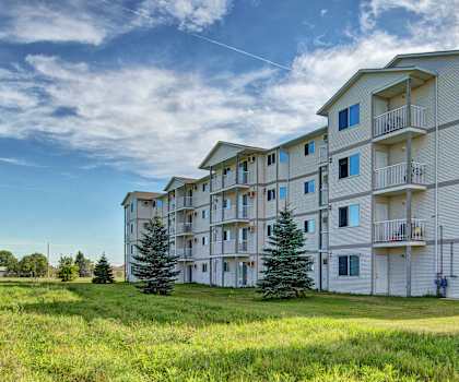 The white exterior of the apartment building surrounded by luscious green grass and bright trees at Villagio Apartment Homes. 