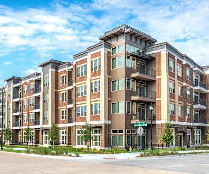 The street exterior view of the red brick, brown, and white apartment complex with windows and a blue sky above at Waterford Market Apartment Homes. 