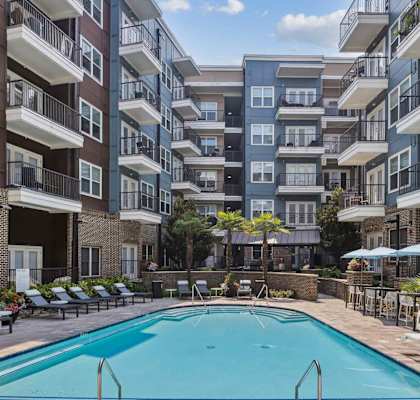 A swimming pool surrounded by lounge chairs and umbrellas in front of apartment buildings.