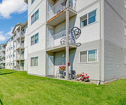 The exterior of the gray apartment buildings with balconies and a grassy area with trees and a fence at Sonora Apartment Homes. 