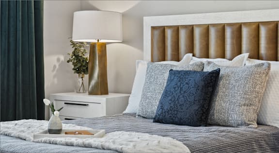 A bed with a white headboard and grey bedding with a gold lamp on a side table.