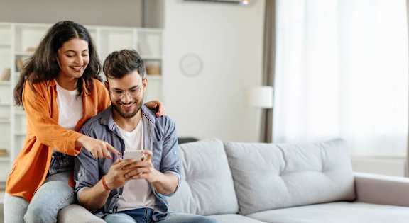 A man and a woman are sitting on a couch, with the woman pointing at the phone the man is holding.