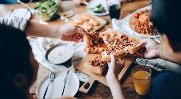 Overhead view of a shared meal on a wood table, with pizza on a wooden board, a salad bowl, and drinks alongside.
