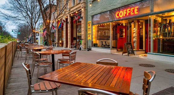 Sidewalk cafe tables and metal chairs outside a lit coffee shop, with string lights strung above the storefronts.
