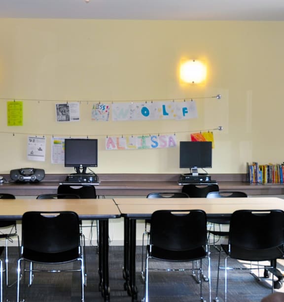 A room with a long table and chairs, a computer desk, and a bulletin board with the word "ALL'S" on it.