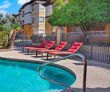 The large, turquoise outdoor pool with red pool chairs, palm trees, with the orange apartments in the background at Sonoma Apartment Homes. 