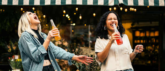 Two women are standing outside a restaurant, one is holding a glass of beer and the other is holding a glass of juice.