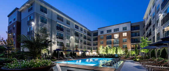 A large building with a pool in the foreground and lights on.