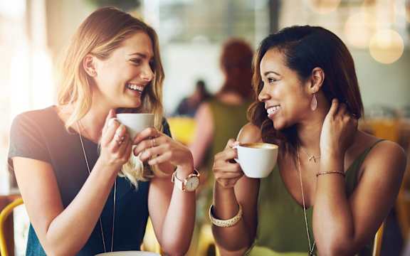 Two women are having a conversation over coffee in a brightly lit café.