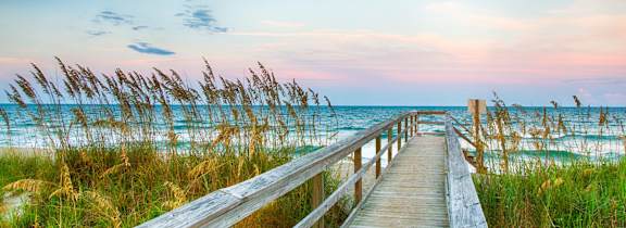 A wooden boardwalk leads to a beach with grasses on either side.