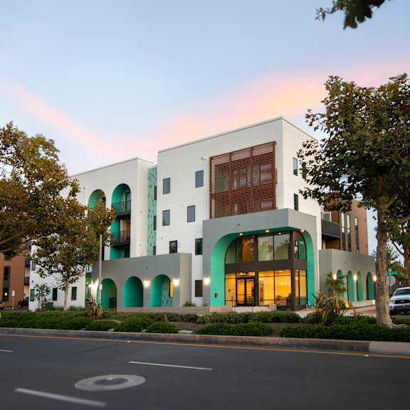 A modern building with a white facade and green accents is surrounded by trees and parked cars.