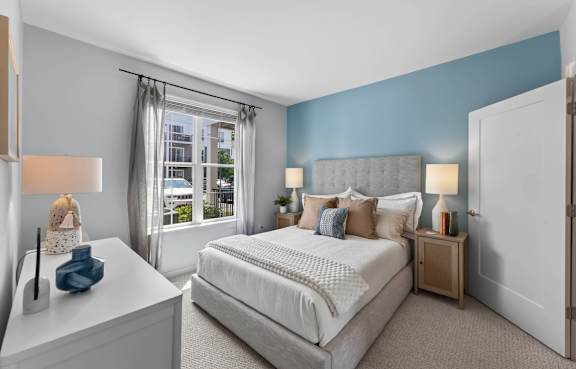 Apartment bedroom with a blue accent wall, upholstered bed, bedside tables, and window.