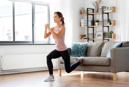 A woman in a pink top and black leggings is doing a lunge exercise in a living room.