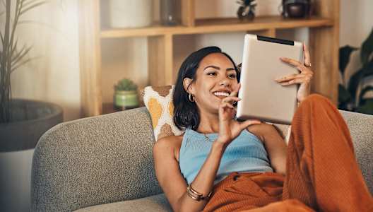 A woman is sitting on a grey couch holding a tablet.
