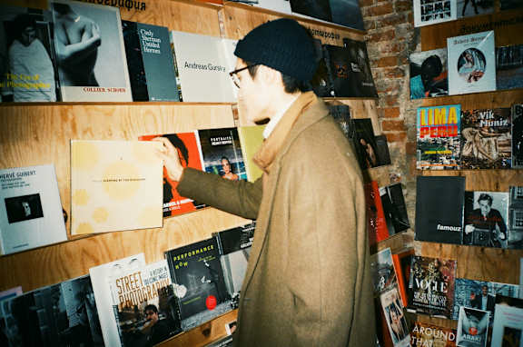 A man in a beige coat and black hat is looking at a book on a wall full of books. at The Fowler Apartments, Boise, ID