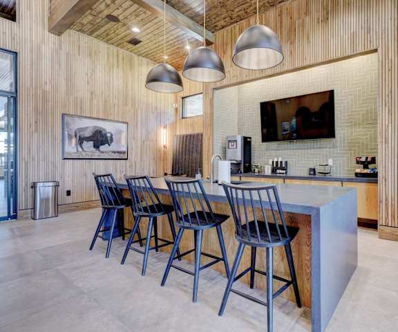 The community kitchen with a TV, a coffee machine, fridge, overhead lights, dark counters, and a kitchen island with barstool chairs at the Encore Apartments. 