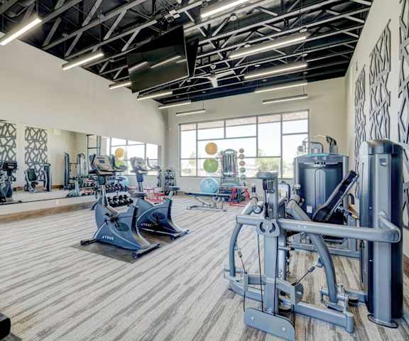 A spacious gym with cardio machines, windows, TVs, and weights at the Encore Apartments. 