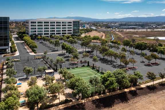 A tennis court is surrounded by trees and a parking lot.
