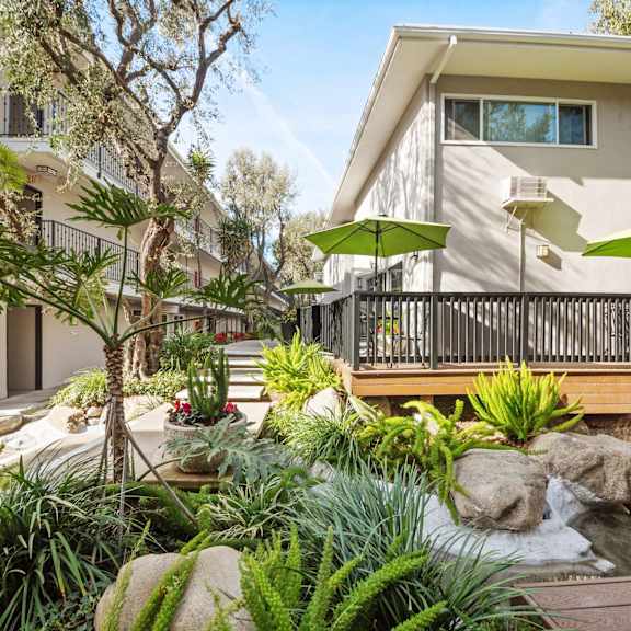 A patio with a table and chairs is surrounded by green plants and trees.